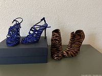 Two pairs of shoes: blue suede heeled sandals and tiger print hide hair booties, displayed on shoe box against a plain wall.