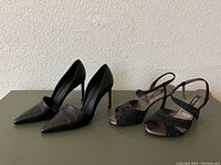 Both pairs of black women's heels displayed on a table against a wall. One pair is pointed-toe leather pumps, the other is black glittery open-toe slingback heels.