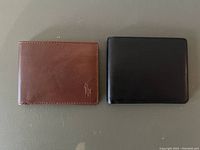 Two men's leather wallets displayed side by side on a flat surface. Left is dark brown with Polo Ralph Lauren logo, right is black.