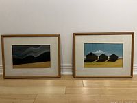 Photo of two framed oil pastel artworks side by side on floor against wall showing landscapes under glass in wood frames.
