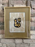 Frontal image of framed abstract artwork hung on wall showing black outlined organic shapes filled with yellow and red on beige textured background.