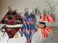 Full view of three swimsuits laid out on a white surface: floral tank-style bikini set, mermaid scale one-piece, and peach/coral bikini set with mint green ties.