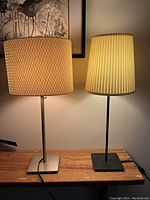 Two table lamps side by side on a wooden surface, one with metal base and textured beige drum shade, the other with black base and cream pleated tapered shade, both lit.