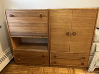 Two matching wood cabinets side by side. Left cabinet has a drop-down door, open middle shelf, and two drawers at the bottom. Right cabinet has two closed doors.