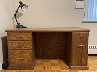 Front view of heavy wood desk with four drawers on left and cabinet door on right, showing wood grain finish and brass handles. Desk holds a black adjustable desk lamp on top.