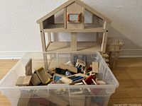 Wooden dollhouse next to plastic container filled with various wooden furniture pieces and dolls.