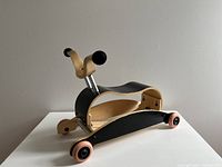 Front angled view showing the curved wooden body with black and natural wood finishes, handlebar grips, and the wheels attached to the black rocker base.