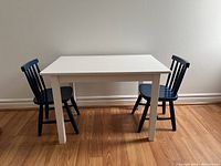 Front view of white painted rectangular wooden table with two child-sized blue wooden chairs on each side on hardwood floor.