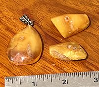 Three pieces of polished Baltic amber: one pendant with a silver bail shaped like a leaf and two loose stones, laid next to a ruler for size reference showing pieces roughly between 1 and 2 inches.