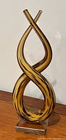 Full view of Murano glass sculpture showing its twisting figure-eight amber form with black streaks mounted on a clear square glass base.