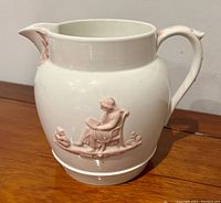 Front side view of white porcelain pitcher with pink relief of a seated woman reading with a dog.