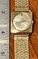 Frontal view showing square gold case, silver dial, baton markers and top portion of woven gold bracelet next to ruler