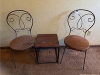 Front view of two metal bistro chairs with velour seats and a wooden metal side table between them, on carpeted floor against wall.