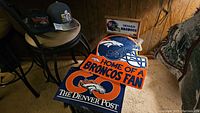 Denver Broncos helmet foam sign, wooden Denver Broncos sign, and Denver Broncos hats on chair and couch