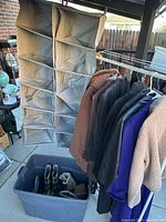 Clothing rack with eight jackets and a large plastic bin containing assorted boots and shoes, two gray fabric hanging closet organizers visible in back.