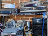 Various vintage electronics including stacked VCRs, CD/DVD players, small space heaters, mini white robotic vacuum, karaoke microphone, and Victor Borge VHS tapes on shelving against brick background.