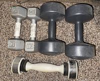 Photo showing two metal hexagonal 10-pound dumbbells, two black plastic 10-pound dumbbells, and one Shake Weight dumbbell on carpet.