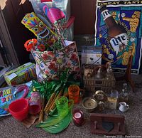 Full lot display showing Hawaiian party decor, plastic cups, bowls, tiki and flamingo decorations, wicker storage, metal Lite Miller beer sign, candles, clocks, shelf, plug-in light, frame holder, and bells.