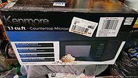 Box of Kenmore countertop microwave oven showing brand, size (1.1 cu.ft), features like six cooking functions, one-touch settings, digital LED display, and defrost options.