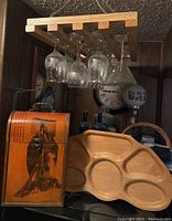 View of 12 etched wine glasses with winter scene designs hanging from light wooden rack. Three clear martini glasses hanging as well including one marked 'BAR'. Below rack is Asian decorative wine bottle holder in orange and black with figure artwork. To right is mid-century modern style wooden serving tray with compartments.