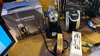 Nespresso VertuoLine coffeemaker in black and chrome with original box, Keurig coffee maker, coffee press, and electric cocktail mixer shown together