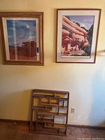 Wide shot showing two framed posters hanging on the wall and mid-century modern wooden wall shelf on the floor beneath them.
