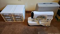 Portable white Necchi Lydia Mk 2 sewing machine with power cord coiled beside a white organizer holding six drawers with sewing notions inside on a carpeted floor.