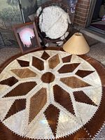 Photo showing large round cowhide rug with geometric star pattern, dreamcatcher, lampshade, and framed painting all arranged on a floor.
