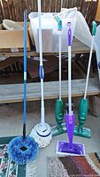 Full view of five cleaning tools: blue broom, white mop, two green Swiffer sweepers, and purple spray mop.