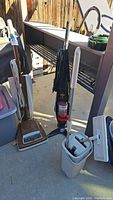 Montgomery Ward upright vacuum, Bissell vacuum, and mop bucket arranged near a shelf outdoors.