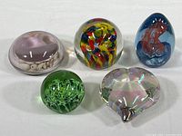 Five glass paperweights arranged closely showing variety in shape, size, and internal designs, including a pink flower-like shape with small shells in one piece