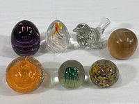 All seven items arranged in two rows showing a mix of glass paperweights and a ceramic bird figurine, detailing various colors and shapes.