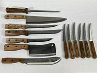 Full set of kitchen cutlery arranged on white cloth, showing the honing steel, large chef's knife with missing tip, butcher cleaver, 2 boning knives, and 6 small serrated knives.