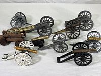 Collection of ten miniature toy cannons in various finishes and colors, arranged on a white surface to show variety and size range.