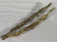 Two vintage brass handles lying parallel on fabric background, showing front side with rhinestone decorations placed centrally on the curved design.