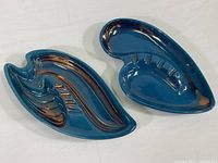Two dark teal blue Mid Century Modern ceramic ashtrays with copper-toned glaze, showing their size, color, and asymmetrical abstract designs