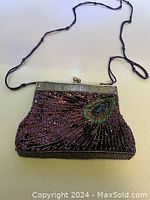 Front view of a purple sequined and beaded small handbag with a peacock feather design, antique bronze metal frame, kiss lock closure, and thin purple cord strap.