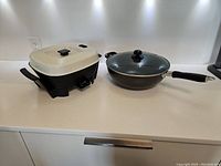 Photo shows Black & Decker electric frying pan with cream-colored lid and black handles next to a Teflon wok with glass lid and black handle, displayed on white countertop.