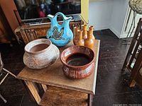 Photo showing the four pottery items together on a wooden table, including the blue pitcher, round pot, brown bowl, and fused vases cluster.
