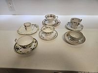 Set of 6 tea cups and saucers of various floral and gold-trimmed patterns displayed on white counter.