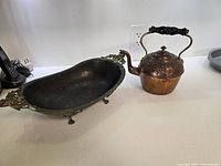 Photo shows copper tea kettle with floral engraving next to oval brass footed bowl with leaf handles