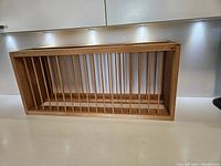 Front view of a rectangular pine wood kitchen plate organizer rack with multiple vertical slats for plate separation, sitting on a light-colored countertop under cabinetry with recessed lighting.