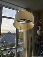 White cylindrical fabric lampshade with visible bulb inside, hanging near a window with curtain and cityscape view outside.