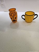 Photo of two yellow Art Deco Czech glass pieces on a white surface: a vase with black bird/tree design and a two-handled cup with black stripes