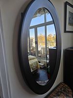 Front view of the large oval mirror showing reflective glass surface and dark rounded frame, mounted on the wall with window reflections visible in the glass.