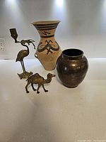 Photo showing four items: a pottery urn with Egyptian designs, a dark brass pot/vase with engravings, a brass camel figurine, and a brass candlestick holder shaped like a crane standing on a turtle.