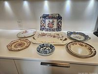 Seven vintage and antique serving pieces displayed on white counter, showing different shapes, colors, and floral patterns to confirm variety and condition.