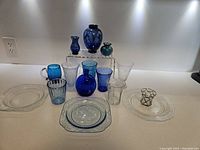 View of entire set of 15 glassware pieces including cups, vases, plates in blue and clear glass