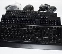 Three black computer keyboards and three computer mice arranged on a white surface, showing keyboards with full key layouts and mice with wired and wireless configurations.