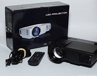 Black LED projector with lens and side input ports, along with remote, power cord, and HDMI cable, next to original box featuring a white projector image.